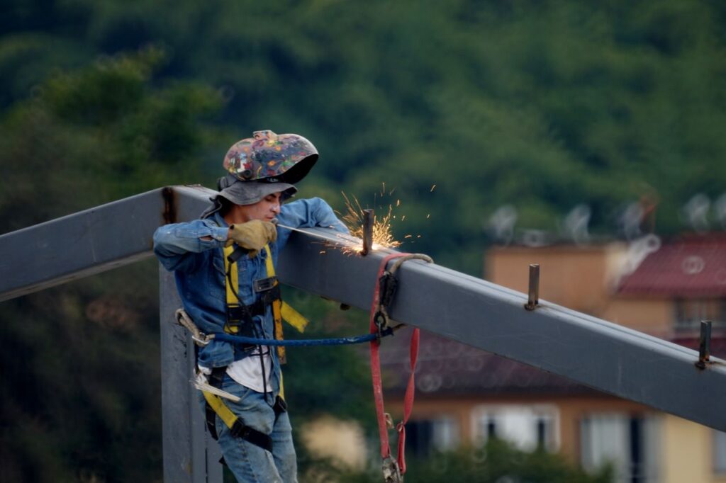 Construction worker welding steel beam on site, wearing safety gear and helmet.